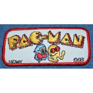 May include: A vintage Pac-Man embroidered patch with a red border. The patch features the iconic Pac-Man character, a ghost, and the text "PAC-MAN" in yellow. The text "MIDWAY" and "©1981" are also visible on the patch.