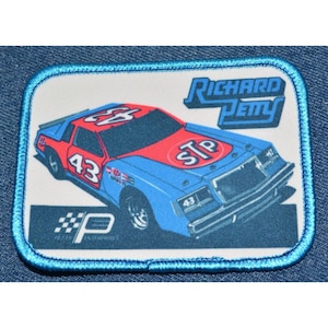 May include: A blue and red race car with the number 43 on the side and the word "STP" on the hood. The car is on a white background with the words "Richard Petty" in black text above the car. The car is surrounded by a blue border.