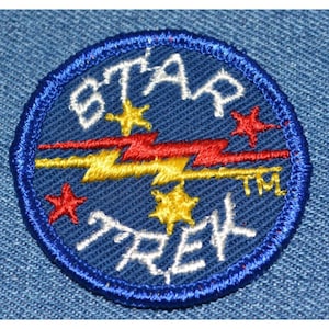 May include: A blue and white embroidered patch with the words "STAR TREK" and a red and yellow lightning bolt design. The patch has a blue border and a white background.