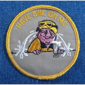 May include: A tan embroidered patch with a yellow border. The patch features a cartoon character wearing goggles and a yellow jacket, holding a glass. The text "HAVE ONE ON ME" is written around the edge of the patch.