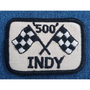 May include: A white embroidered patch with black stitching. The patch features two crossed checkered flags and the text "500 INDY".
