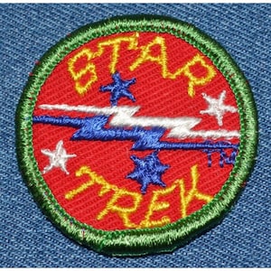 May include: A red, green, and white embroidered patch with the words "STAR TREK" in yellow lettering. The patch features a lightning bolt design and stars.