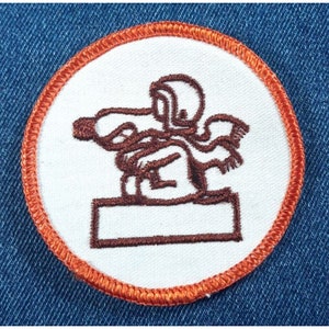 May include: A brown embroidered patch with an orange border. The patch features a cartoon dog wearing a helmet and standing on a platform.