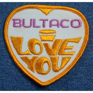 May include: A white heart-shaped patch with an orange border. The patch has the text "BULTACO" in pink lettering and "LOVE YOU" in yellow lettering.