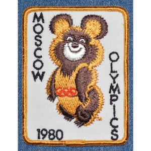 May include: Embroidered patch featuring the Moscow Olympics bear mascot. The bear is brown and gold, with a smiling face and Olympic rings on its chest. The patch includes the text "MOSCOW OLYMPICS 1980" in black lettering.