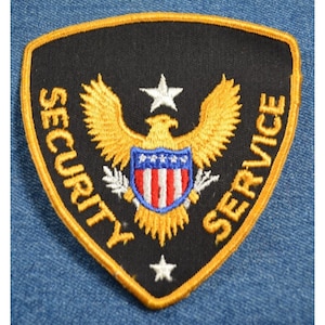 May include: Black embroidered patch with a gold outline. The patch features a gold eagle with a shield in its talons. The shield has red and white stripes and a blue background. The eagle is holding a star above its head. The words "SECURITY SERVICE" are written in gold around the eagle.