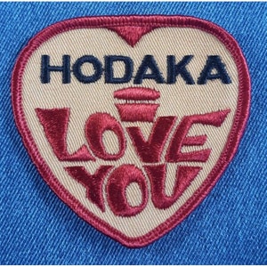 May include: A tan heart-shaped patch with the text "HODAKA LOVE YOU" in black and red embroidery. The word "LOVE" is in a stylized font and the word "YOU" is in a smaller font.