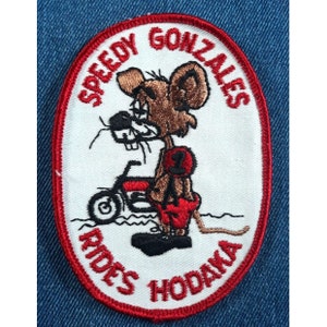 May include: A white oval embroidered patch with red stitching. The patch features a cartoon mouse riding a motorcycle with the text "Speedy Gonzales Rides Hodaka" around the edge.