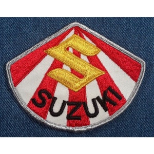 May include: A red, white, and gold embroidered patch with the word "SUZUKI" in black lettering. The patch features a stylized "S" in gold within a red and white sunburst design.