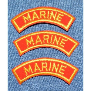 May include: Three red embroidered patches with the word "MARINE" in gold lettering. Each patch has a curved shape and a gold border. The patches are displayed on a blue denim background.