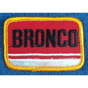 May include: A red and white embroidered patch with a yellow border. The patch says "BRONCO" in black lettering.