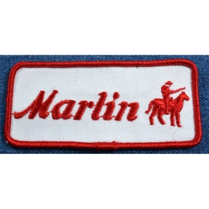 May include: Red embroidered patch with the word "Marlin" in a cursive font and a red silhouette of a cowboy riding a horse to the right of the word.