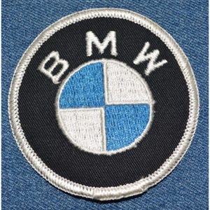 May include: A black and white embroidered patch with the BMW logo. The logo is a white circle with a blue and white propeller design inside. The letters BMW are embroidered in white around the circle.