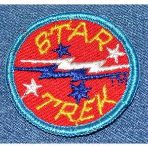 May include: A red and blue embroidered patch with the words "STAR TREK" in yellow lettering. The patch features a white lightning bolt design with blue and white stars.