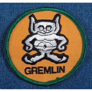 May include: A round, orange patch with a green border. The patch features a white embroidered cartoon character with the word "GREMLIN" in black below it.