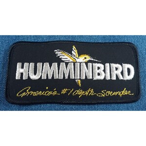 May include: A black embroidered patch with a white hummingbird and the text "Humminbird" in white. Below the hummingbird is the text "America's #1 Depth Sounder" in gold.