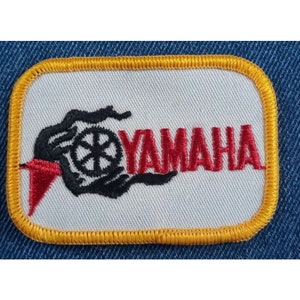 May include: A white embroidered patch with a yellow border. The patch features a black and white stylized image of a motorcycle engine with the word "YAMAHA" in red lettering.