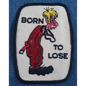 May include: A white embroidered patch with a cartoon figure in a red suit and the text "BORN TO LOSE".