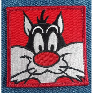 May include: A red embroidered patch featuring a cartoon cat with black fur, white eyes, and a red nose. The cat is looking to the right.