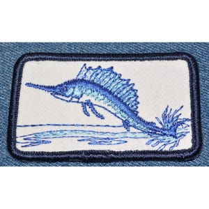 May include: A blue embroidered patch of a marlin jumping out of the water. The patch is on a white background with a blue border.