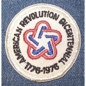 Puede incluir: Un parche circular con el texto "AMERICAN REVOLUTION BICENTENNIAL 1776-1976" bordado en negro. El parche presenta un fondo blanco con un diseño en forma de estrella roja, blanca y azul en el centro. El parche está cosido sobre una tela vaquera azul.