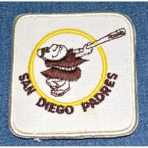 May include: Square white patch with a baseball-themed graphic. The patch features a brown cartoon figure swinging a baseball bat inside a yellow circle. The words "SAN DIEGO PADRES" are embroidered in brown around the circle.