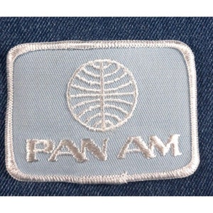 May include: A light blue embroidered patch with a white outline. The patch features a white globe with lines radiating from the center and the text "PAN AM" in white.