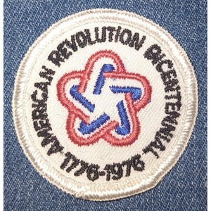 以下が含まれることがあります： 黒字で「AMERICAN REVOLUTION BICENTENNIAL 1776-1976」と書かれた円形のパッチ。パッチは白い背景に赤、白、青の星型のデザインが中央にあります。パッチは青色のデニム生地に縫い付けられています。