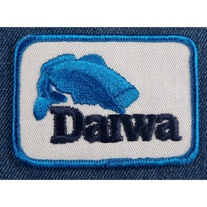 May include: A white embroidered patch with a blue fish and the word "Daiwa" in black lettering.