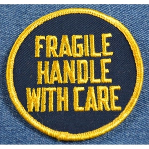May include: A round, navy blue patch with a gold border and the text "FRAGILE HANDLE WITH CARE" embroidered in gold.