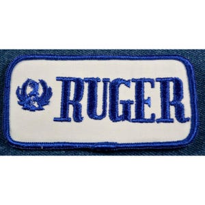 May include: A white rectangular patch with blue embroidered edging. The patch features a blue stylized eagle logo and the word "RUGER" in blue embroidered lettering.