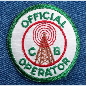 May include: A white embroidered patch with a green border. The patch says "Official CB Operator" in a circle around a red circle with a brown antenna tower and the letters "CB" on either side of the tower.