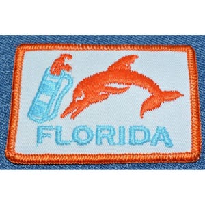May include: A white embroidered patch with an orange dolphin and a blue and white striped bottle. The text "FLORIDA" is embroidered in blue below the image.