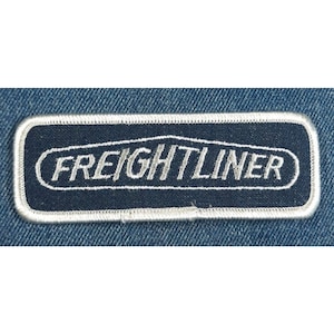May include: A white embroidered patch with a navy blue background. The patch features the text "FREIGHTLINER" in a white oval.
