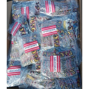 以下が含まれることがあります： プラスチックで包装された多数のパッチが詰まった箱。各パッチは「PAC-MAN」と「NOVAG」の文字が入ったパックマンのデザインが特徴です。パッチは黄色、青、赤など様々な色があり、「SPORTSTRIPE」のラベルでパッケージされています。