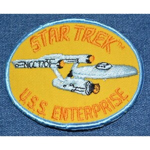 May include: A yellow and blue embroidered patch with the text "STAR TREK" and "U.S.S. ENTERPRISE" surrounding a white and blue spaceship.