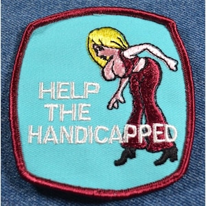 May include: A turquoise blue embroidered patch with a red border. The patch features a cartoon woman with blonde hair wearing a red top and pants. The text "HELP THE HANDICAPPED" is embroidered in white.
