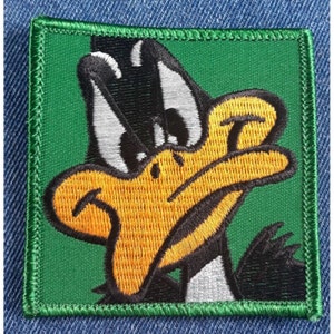 May include: Embroidered patch featuring a cartoon duck with a black and white head, yellow beak, and black body. The duck has a grumpy expression and is set against a green background.