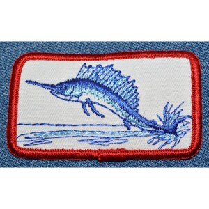 May include: A blue embroidered patch of a marlin jumping out of the water. The patch has a red border and is on a white background.