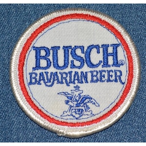 May include: A round patch with the text "BUSCH BAVARIAN BEER" in blue, surrounded by a red and white border. The patch features a blue emblem at the bottom. The patch is on a blue denim background.