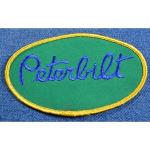 May include: A green oval-shaped patch with a gold border. The word "Peterbilt" is embroidered in blue on the patch.