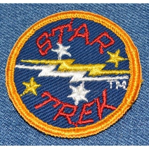 May include: A blue and orange embroidered patch with the words "STAR TREK" and a lightning bolt design. The patch has five white stars and two yellow stars.