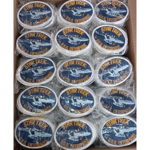 Puede incluir: Una colección de parches ovalados de Star Trek. Cada parche presenta las palabras "STAR TREK" en letras doradas sobre una imagen de la U.S.S. Enterprise y las palabras "U.S.S. ENTERPRISE" en letras blancas.