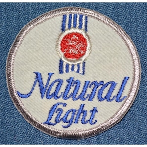 May include: A circular patch with the words "Natural Light" embroidered in blue script. The patch features a red and white emblem at the top and is bordered with a silver thread. The patch is on a denim background.