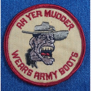 May include: A beige embroidered patch with a red border. The patch features a cartoon image of a person wearing a hat and a menacing expression. The text "Army Boots" and "Ryer Mudder" are embroidered around the image.