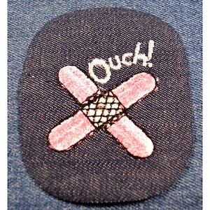 May include: A pink embroidered bandage with a black mesh center and the word "Ouch!" in white embroidery on a dark blue background.