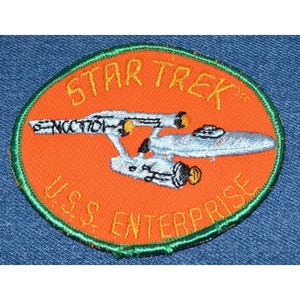 May include: A round orange patch with a green border. The patch features a white spaceship with yellow details and the text "STAR TREK" and "U.S.S. ENTERPRISE" in yellow.