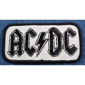 May include: Black and white embroidered patch with the band name "AC/DC" in a bold, stylized font.