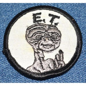 May include: A white embroidered patch with a black border featuring a gray alien with the text "E.T." above it.