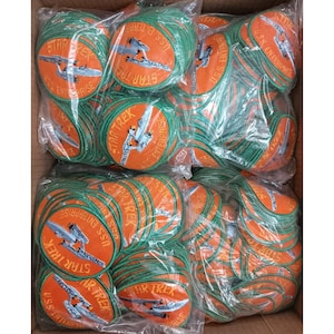 Puede incluir: Una caja llena de parches redondos, naranjas y verdes de Star Trek. Cada parche presenta las palabras "STAR TREK" y "U.S.S. ENTERPRISE" alrededor de un gráfico de una nave espacial. Los parches están empaquetados en plástico transparente.